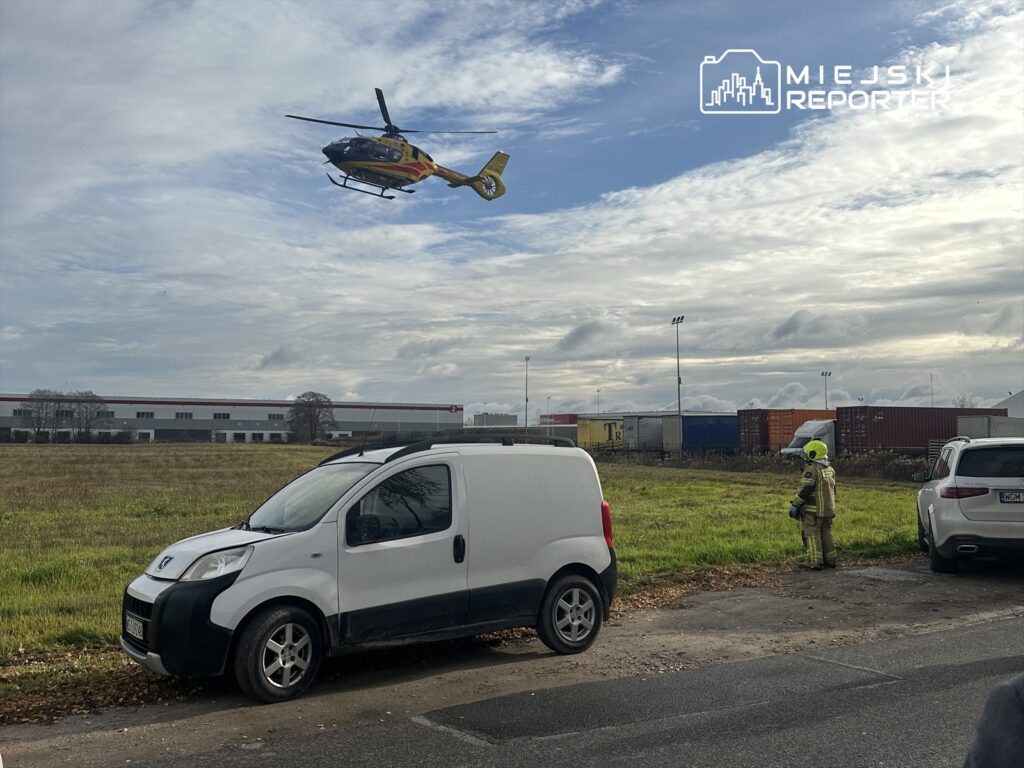 Helikopter ratunkowy przelatuje nad terenem z zieloną trawą, obok stoi strażak w hełmie ochronnym.