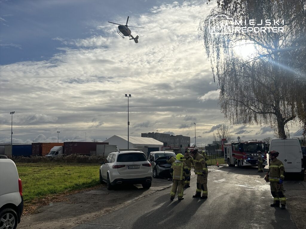 Grupa strażaków w mundurach bada miejsce wypadku drogowego, obok stoi wóz strażacki, nad nimi przelatuje helikopter.