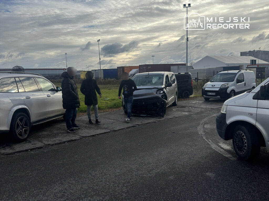 Grupa ludzi stoi obok uszkodzonego pojazdu na ulicy, w tle widać kontenery i hale.