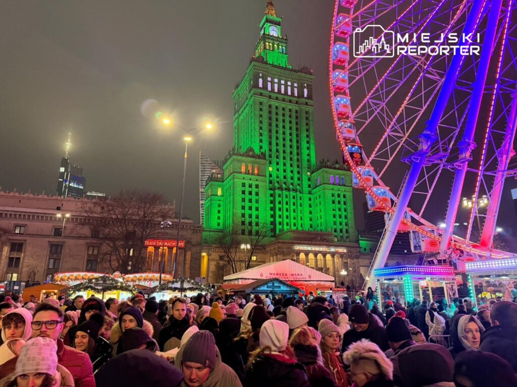 Tłumy ludzi biorące udział w jarmarku pod oświetlonym zielonym Pałacem Kultury, w tle diabelski młyn.