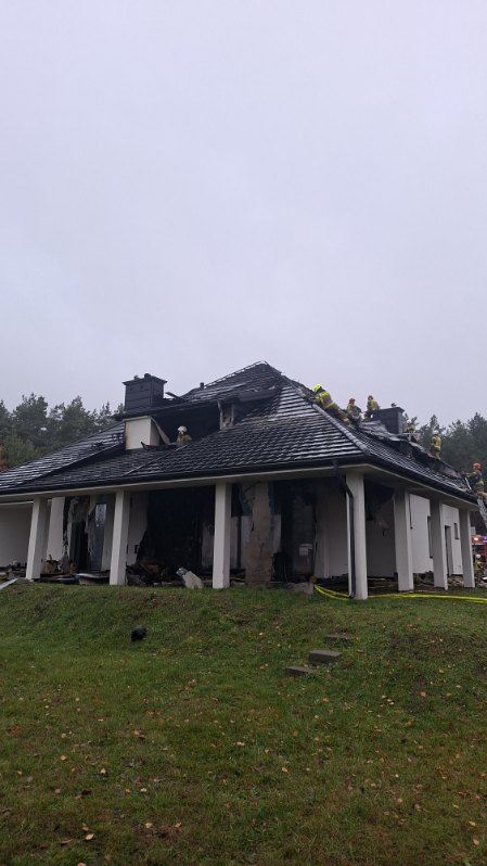 Strażacy pracują nad ugaszeniem pożaru na dachu nowoczesnego domu w otoczeniu lasu.