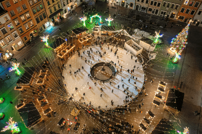 Tłum ludzi tańczących na lodowisku w centrum miasta, otoczonym świątecznymi dekoracjami i oświetleniem.