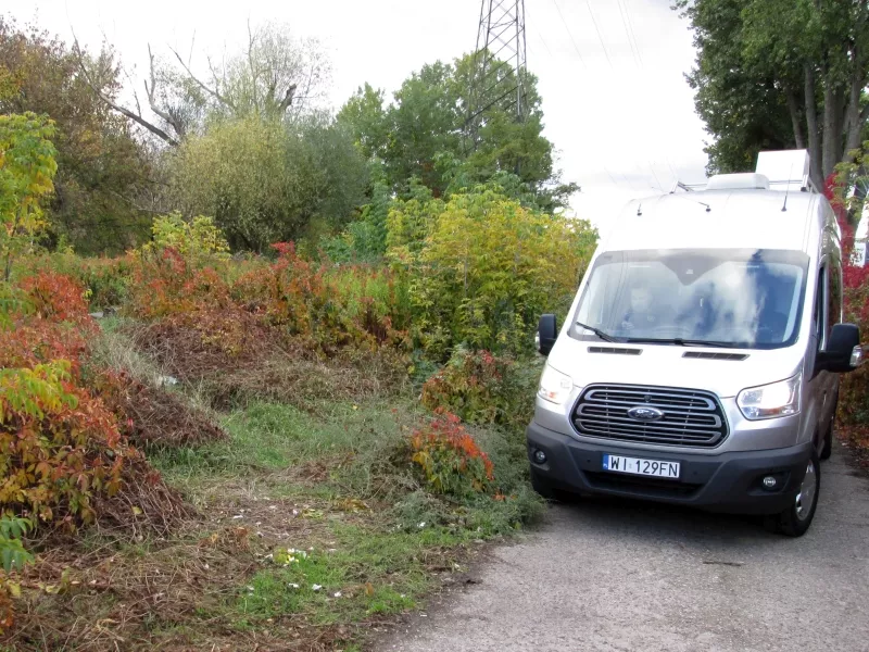 Samochód dostawczy zaparkowany na wąskiej drodze otoczonej krzewami w jesiennych kolorach.
