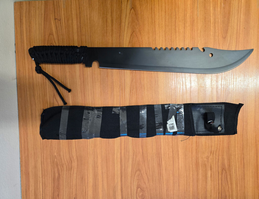 Nóż o dużym ostrzu leży na drewnianym blacie obok czarnej osłony z materiału.