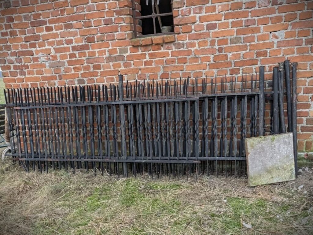 Metalowy płot z ostrymi końcami leży na trawie obok ceglanej ściany budynku.