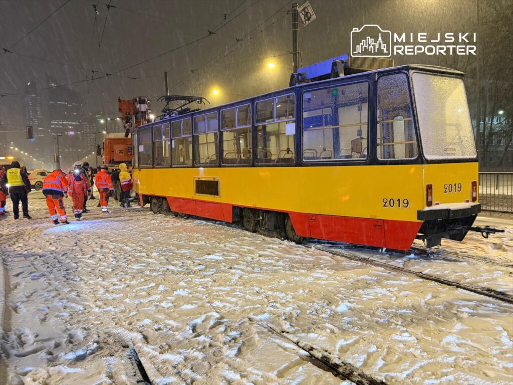 Zespół pracowników w pomarańczowych uniformach naprawia tramwaj na zaśnieżonej ulicy w nocy.