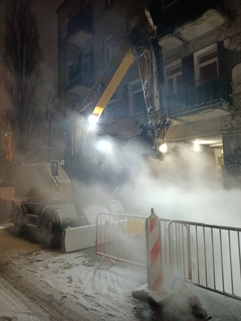 Maszyna budowlana z wysięgnikiem pracuje na zaśnieżonej ulicy, otoczona mgłą i barierkami.