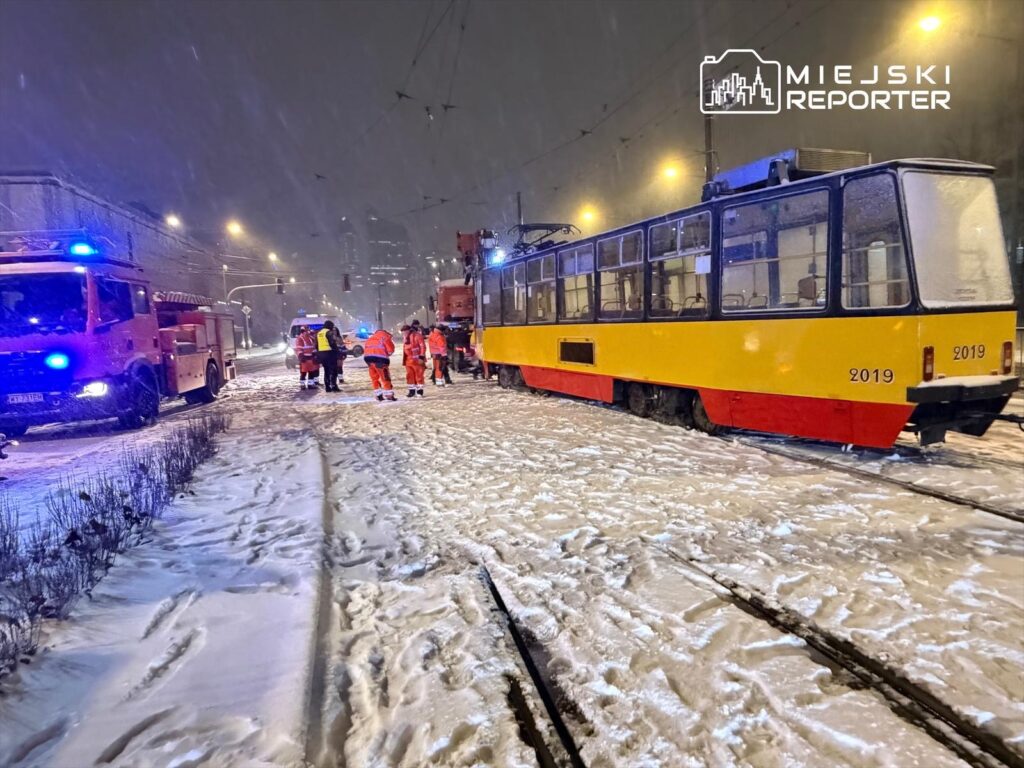 Ratownicy w odblaskowych strojach pracują obok tramwaju na zaśnieżonej ulicy, a w tle widać woz strażacki.
