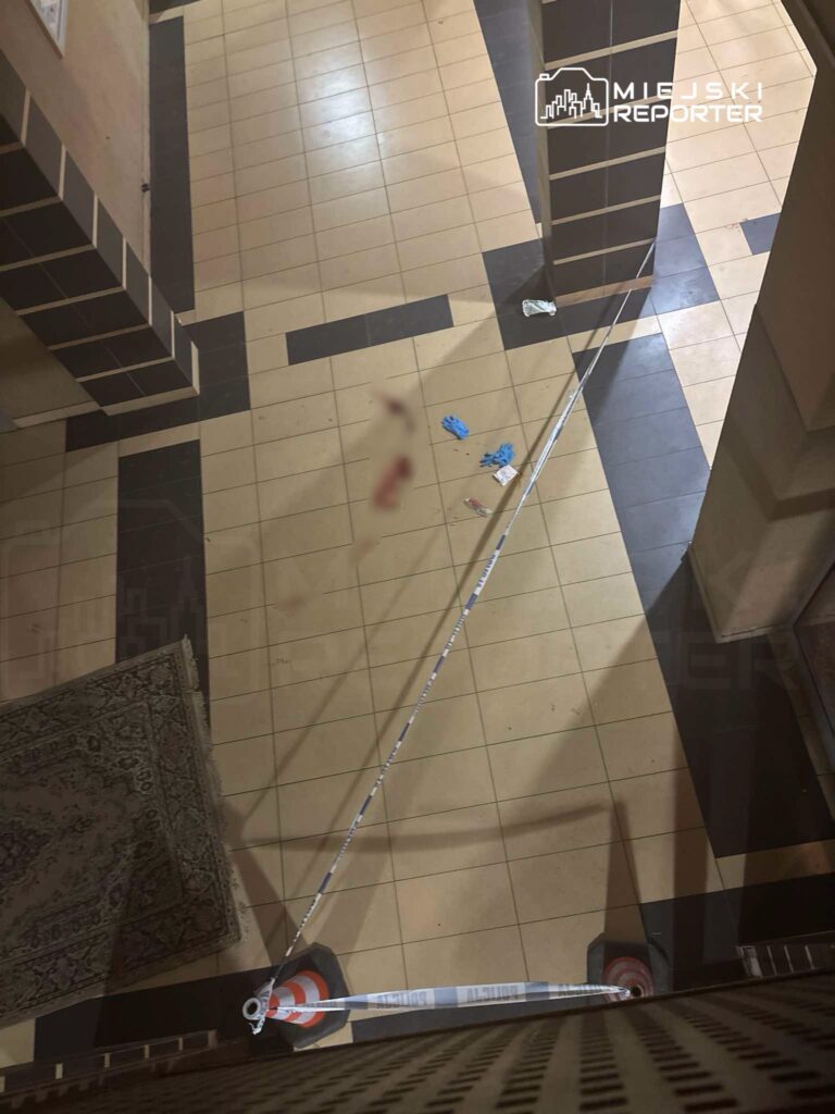 Korytarz w budynku z plamą na podłodze oraz niebieskimi rękawiczkami, zabezpieczony taśmą policyjną.