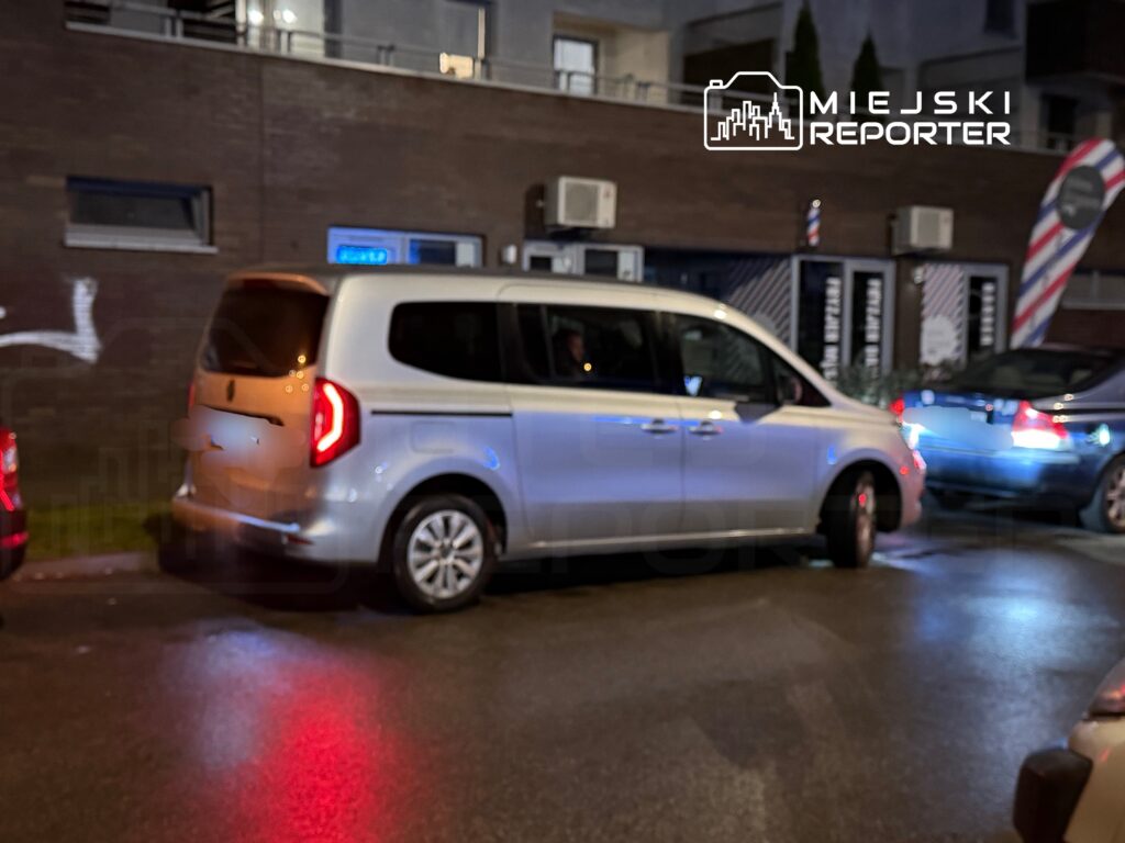 Srebrny van zaparkowany obok ciemnego budynku, w tle widoczne oświetlenie ulicy i inne samochody.