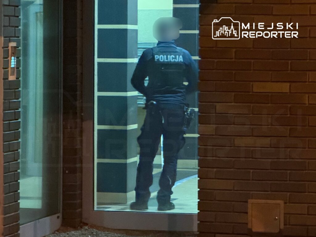 Funkcjonariusz policji stoi przy drzwiach szklanych w nowoczesnym budynku, obserwując otoczenie.