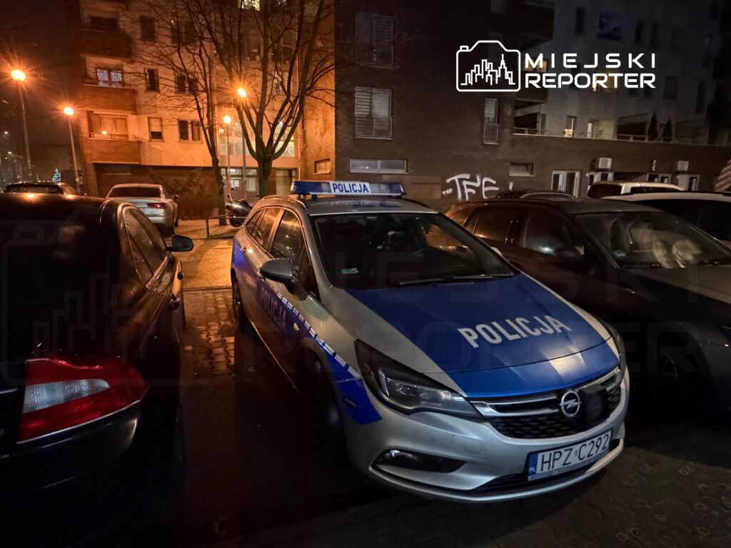 Policyjny radiowóz zaparkowany w ciemnej ulicy, obok innych samochodów, w nocy.