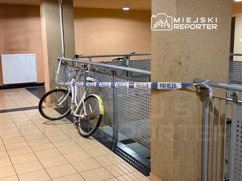 Biały rower przypięty do balustrady, zabezpieczony taśmą policyjną w przestrzeni publicznej.