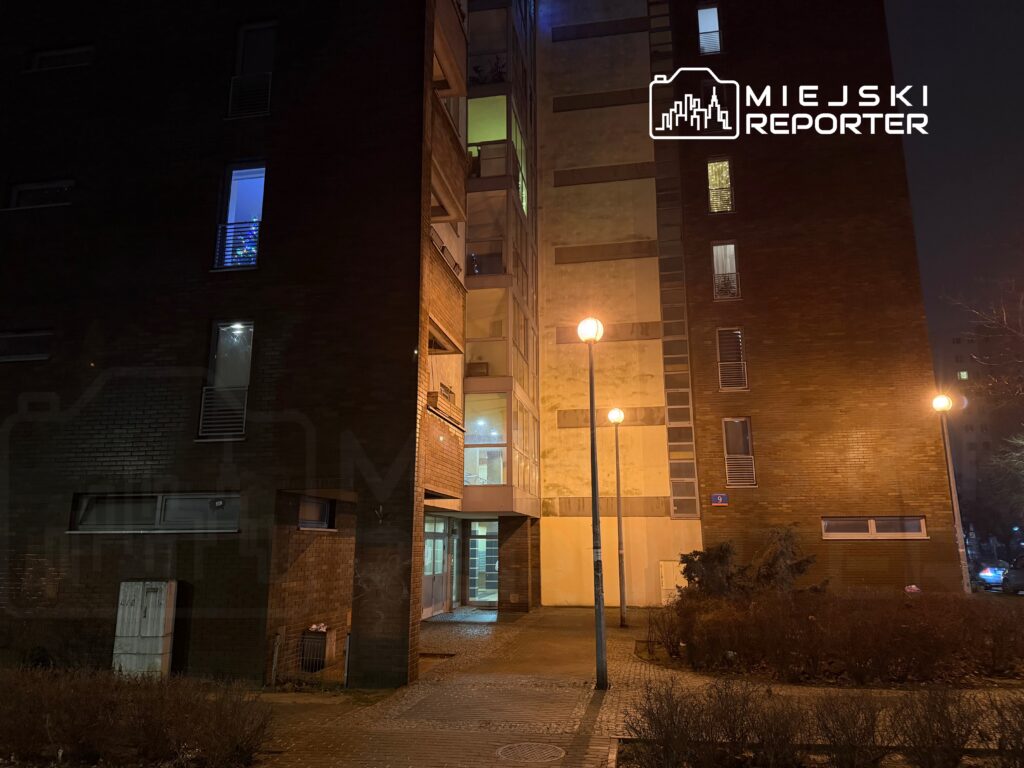 Nowoczesny budynek mieszkalny o ceglanej elewacji, oświetlony lampami ulicznymi, w nocy z widocznymi zapalonymi oknami.
