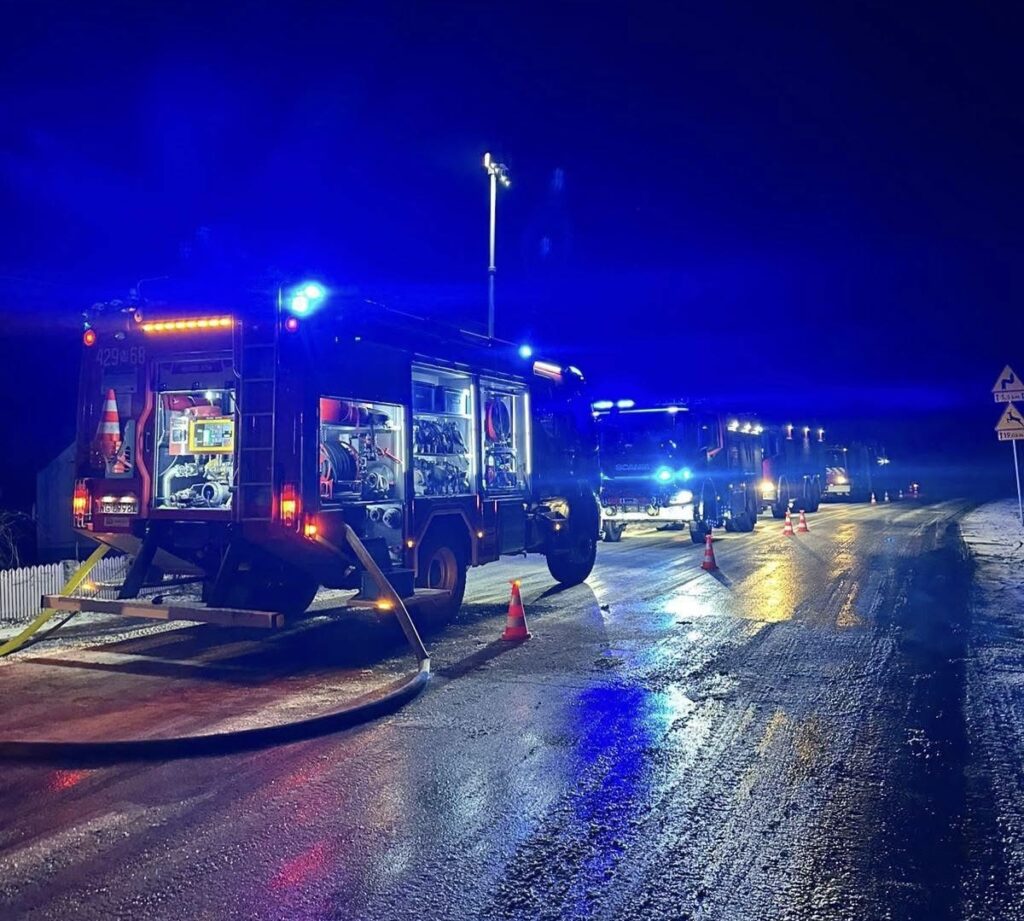 W nocy na drodze zatrzymane są wozy strażackie z włączonymi sygnalizacjami świetlnymi, obok leży wąż strażacki.