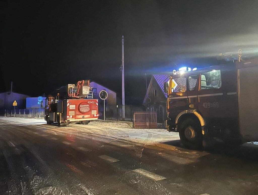 Straż pożarna z włączonymi sygnałami świetlnymi stoi na ośnieżonej drodze w nocy, obok budynków mieszkalnych.