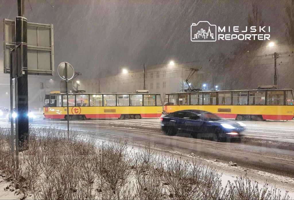 Tramwaje w żółto-czerwonych barwach zatrzymane w śnieżnej scenerii, obok przejeżdżający samochód.