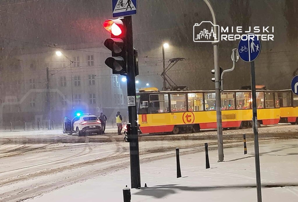 Tramwaj w kolorach żółto-czerwonych stoi na przystanku, obok policyjny samochód z włączonymi sygnałami świetlnymi w śnieżnej scenerii.