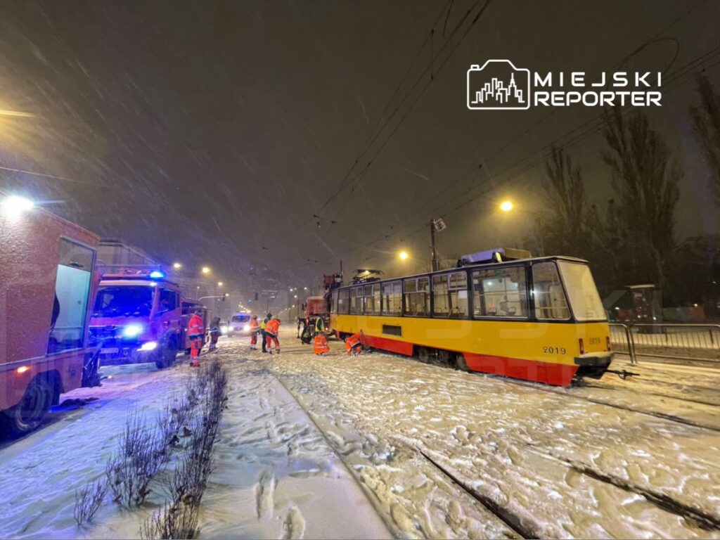 Ratownicy w pomarańczowych kurtkach pracują przy wykolejonym tramwaju na zaśnieżonej ulicy w nocy.