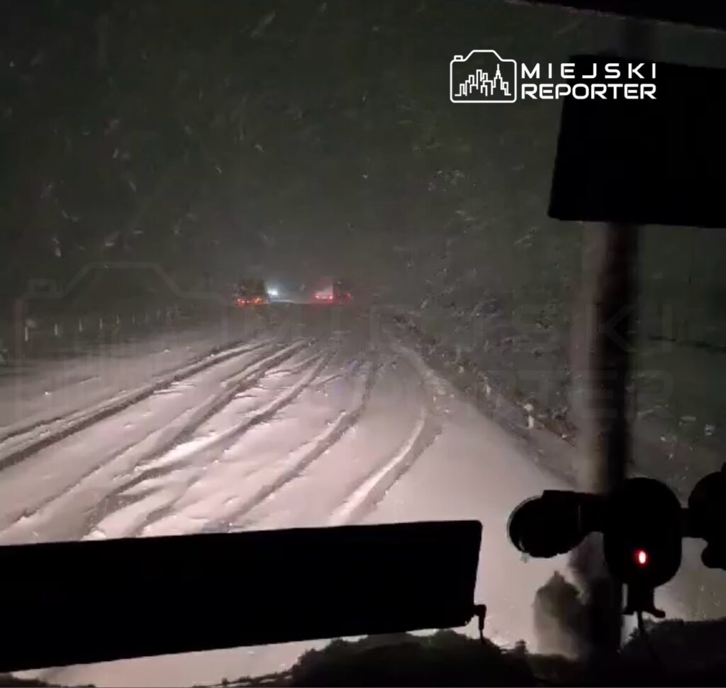 Widok z wnętrza pojazdu na zaśnieżoną drogę, z widocznymi śladami opon i słabym oświetleniem w tle.