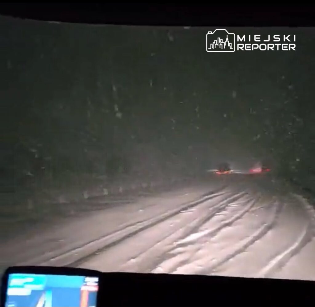 Widok z wnętrza pojazdu na zasypaną śniegiem drogę, z widocznymi śladami kół i padającym śniegiem.