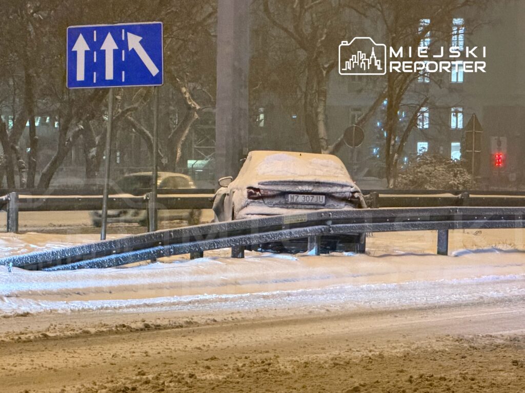 Samochód przykryty śniegiem zaparkowany przy barierce, w tle znaki drogowe w śnieżnej scenerii.