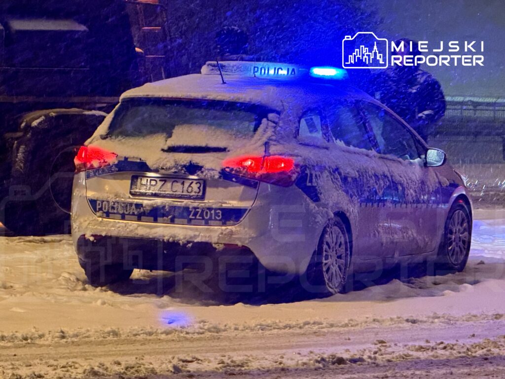 Policyjny samochód zaparkowany na zaśnieżonej drodze, z włączonymi niebieskimi światłami.