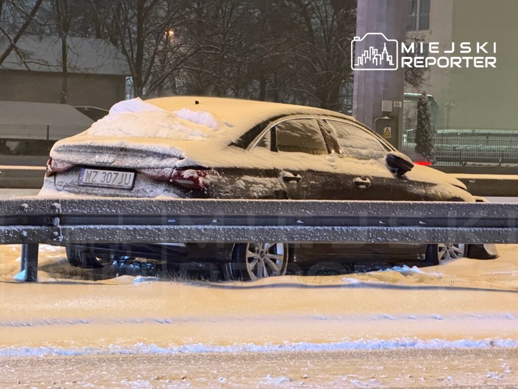 Czarny samochód osobowy pokryty śniegiem, zaparkowany obok bariery na ośnieżonej drodze.