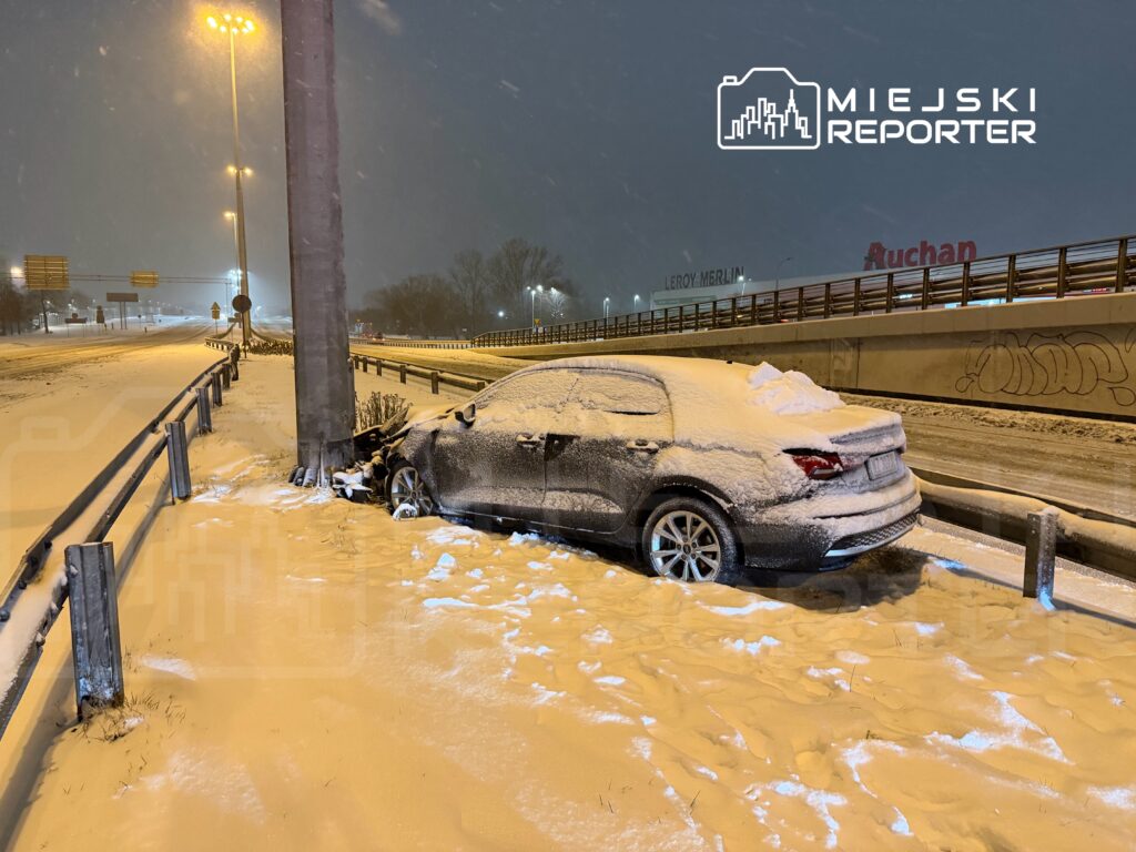 Samochód zaparkowany obok słupa oświetleniowego, pokryty śniegiem, na zaśnieżonej drodze w nocy.
