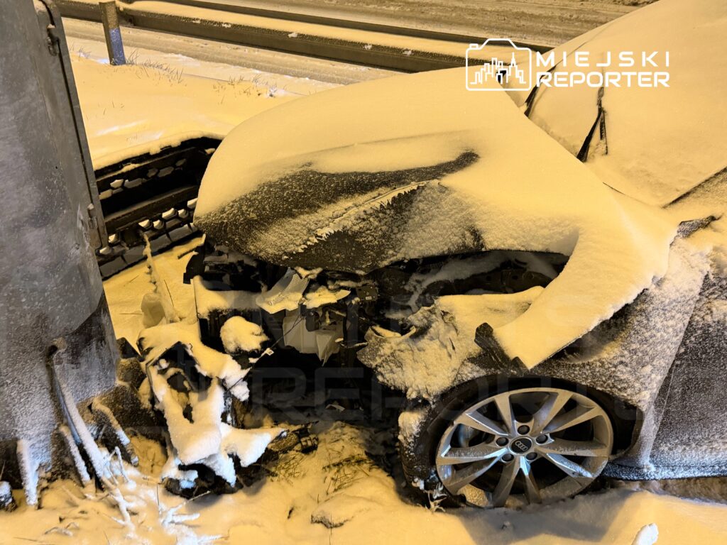 Zniszczony samochód osobowy uderzony w latarnię, pokryty śniegiem, na ośnieżonej drodze przy krawędzi jezdni.