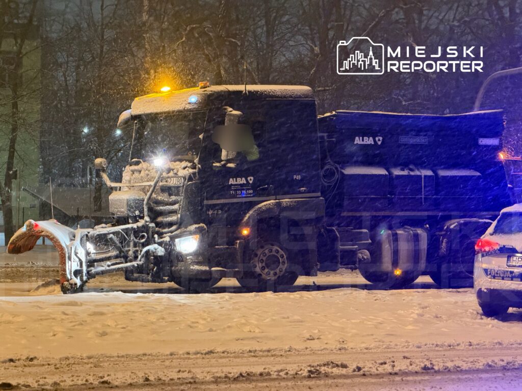 Pojazd z pługiem odśnieżającym pracuje na zaśnieżonej ulicy w nocy, oświetlony przez latarnie.