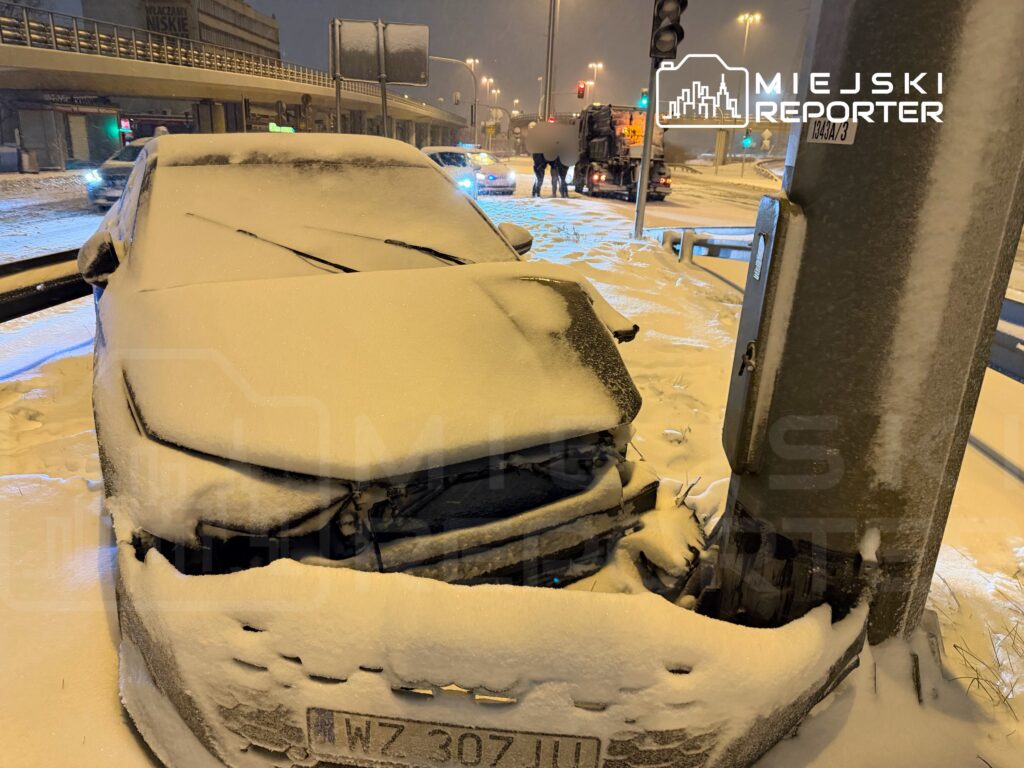 Zasypany śniegiem samochód osobowy zaparkowany na poboczu w nocy, w tle widać ruch uliczny i sygnalizację świetlną.