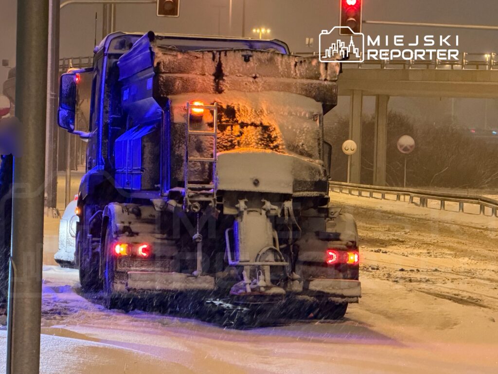 Pojazd ciężarowy z pługiem na zaśnieżonej drodze, w tle sygnalizacja świetlna i oznakowanie drogowe.
