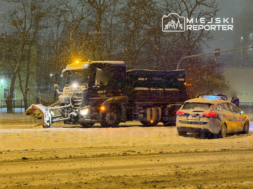 Pojazd do odśnieżania pracujący na zaśnieżonej ulicy, obok radiowóz policyjny z włączonymi światłami.