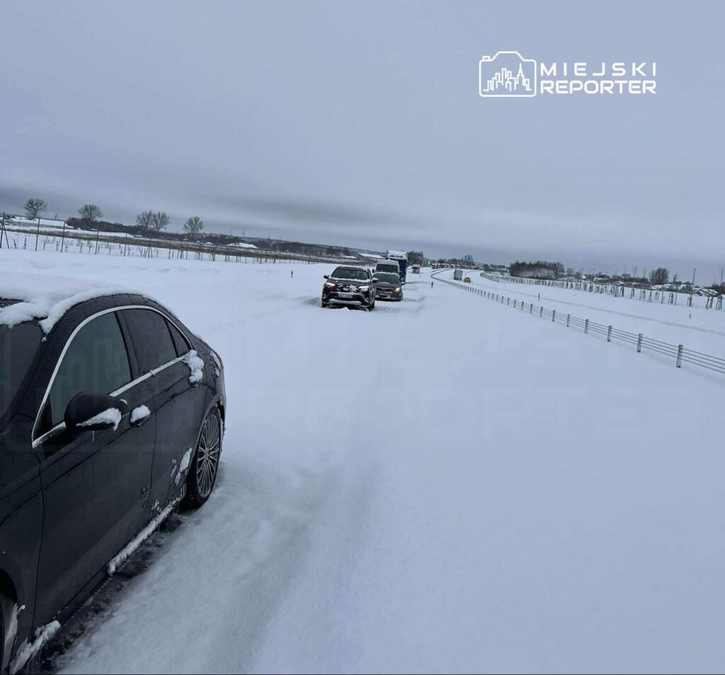 Cztery samochody stoją na ośnieżonej drodze, otoczonej białym śniegiem i szarym niebem.