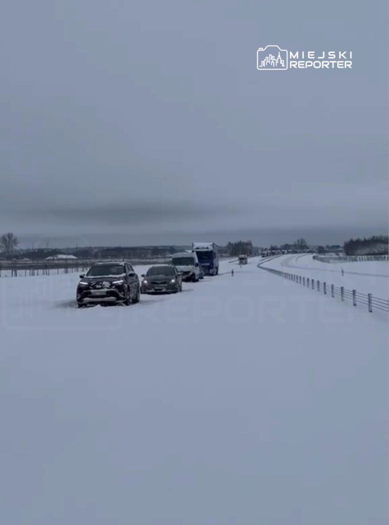 Cztery samochody poruszające się po zaśnieżonej drodze w zimowym krajobrazie. W tle widoczne są zarysy drzew i ogrodzenia.