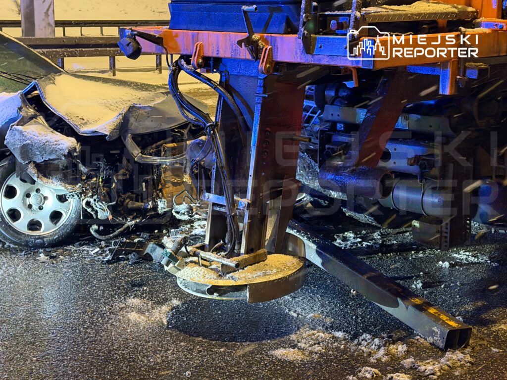 Uszkodzony samochód osobowy z wgniecioną tylną częścią stoi na zaśnieżonej jezdni obok pojazdu służb ratunkowych.