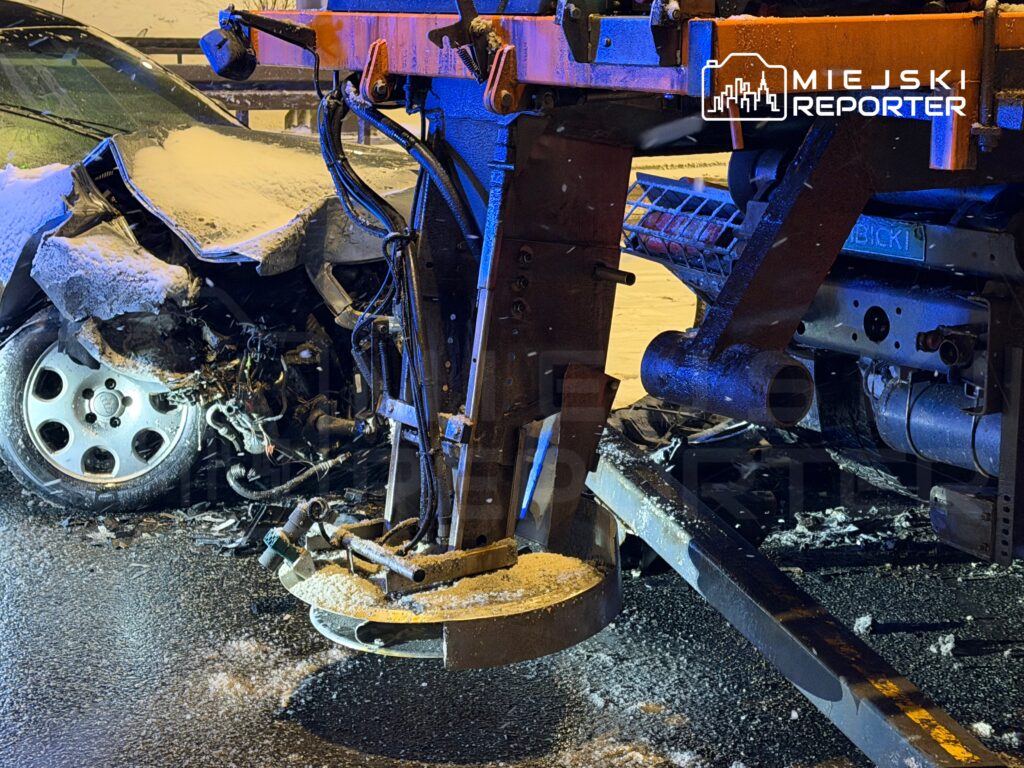 Uszkodzony samochód osobowy z wgniecioną karoserią stoi obok maszyny do odśnieżania na zaśnieżonej drodze.