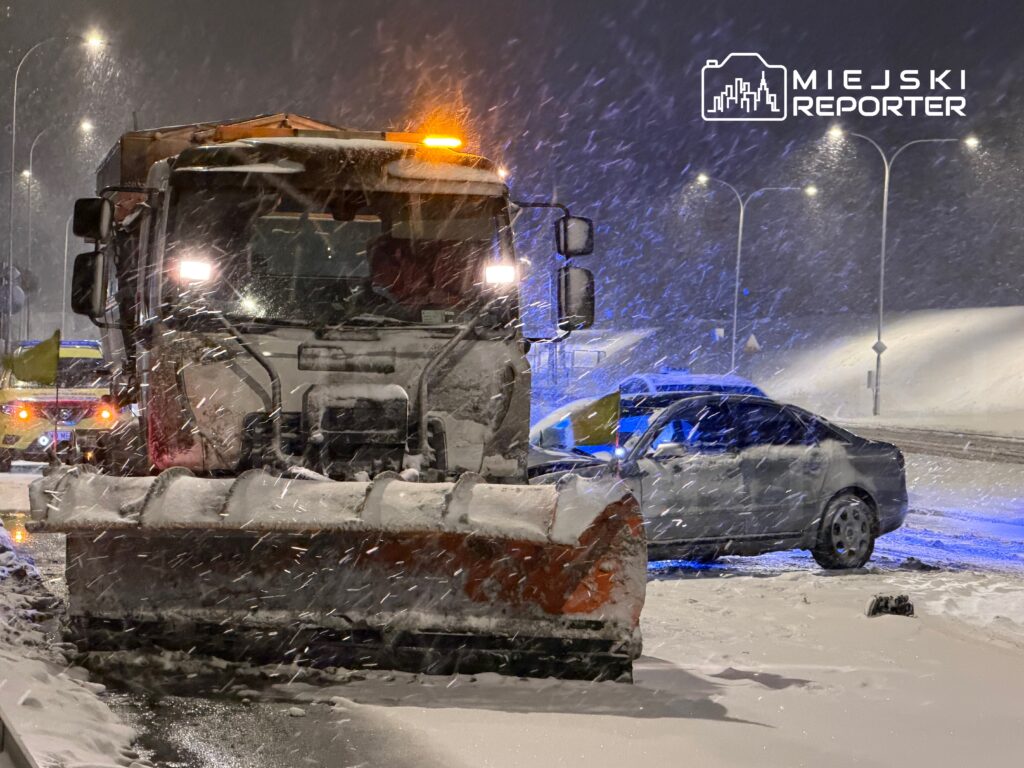 Pojazd drogowy z pługiem śnieżnym odśnieża zaśnieżoną drogę w nocy podczas intensywnego opadu śniegu.