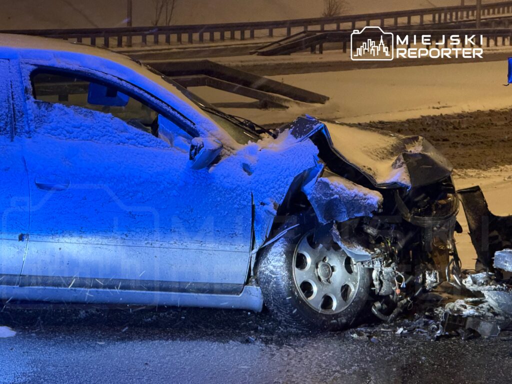 Zniszczony samochód osobowy z poważnymi uszkodzeniami przód w zaśnieżonym otoczeniu nocnym, oświetlony niebieskimi światłami.