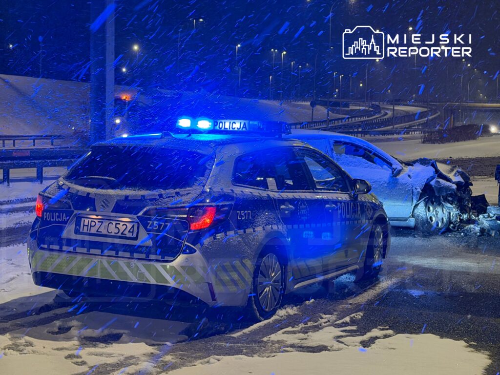 Policyjny radiowóz z niebieskimi światłami zaparkowany obok uszkodzonego samochodu na zaśnieżonej drodze w nocy.