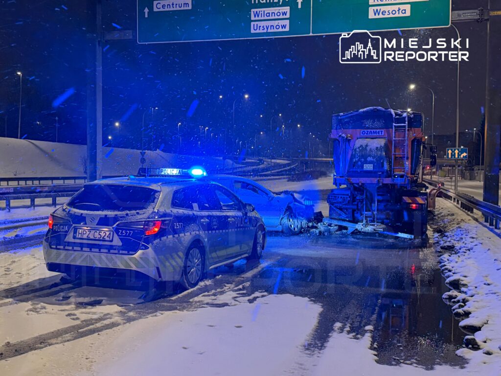 Policyjny radiowóz z włączonymi światłami na ośnieżonej drodze, obok uszkodzonego samochodu i pojazdu do odśnieżania.