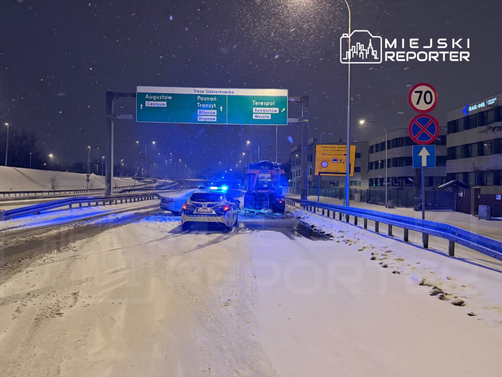 Radiowóz policyjny z włączonymi sygnałami świetlnymi stoi na zaśnieżonej drodze, obok znaku drogowym kierującym do Poznania.