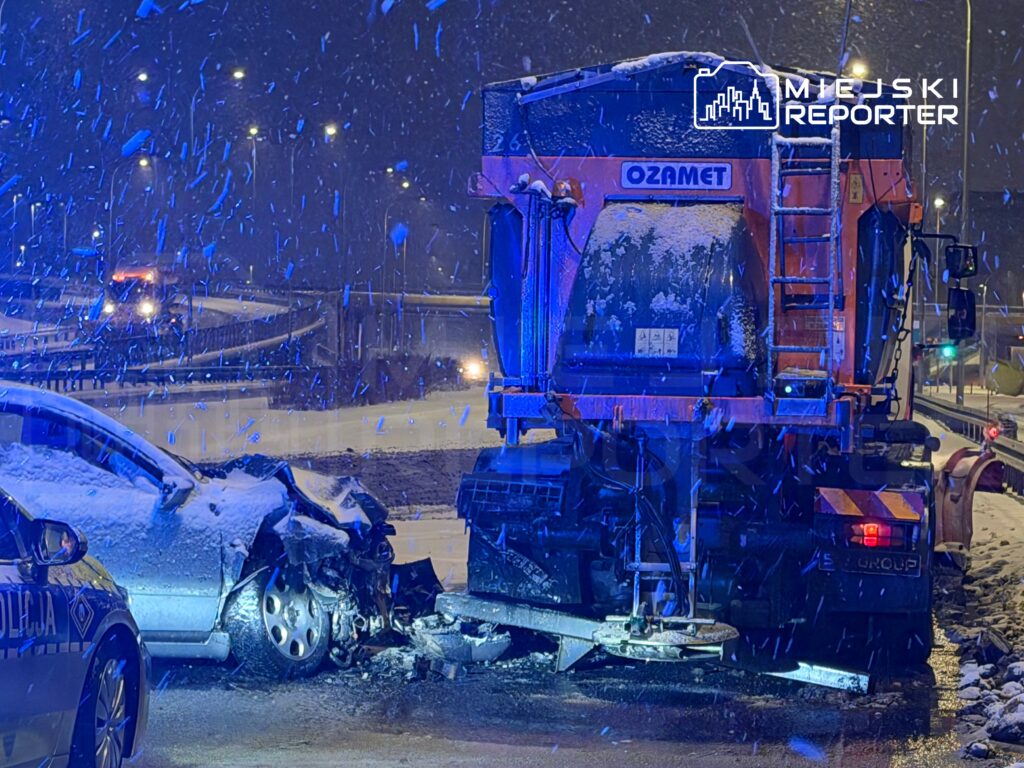 Zderzenie samochodu osobowego z pojazdem drogowym w warunkach śnieżnych, oświetlone przez niebieskie światła policyjne.