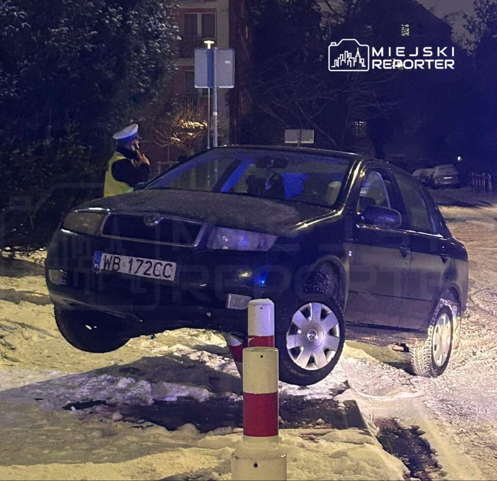 Czarny samochód osobowy unosi się nad betonowym słupkiem w ośnieżonym otoczeniu, obok stoi policjant w kamizelce.