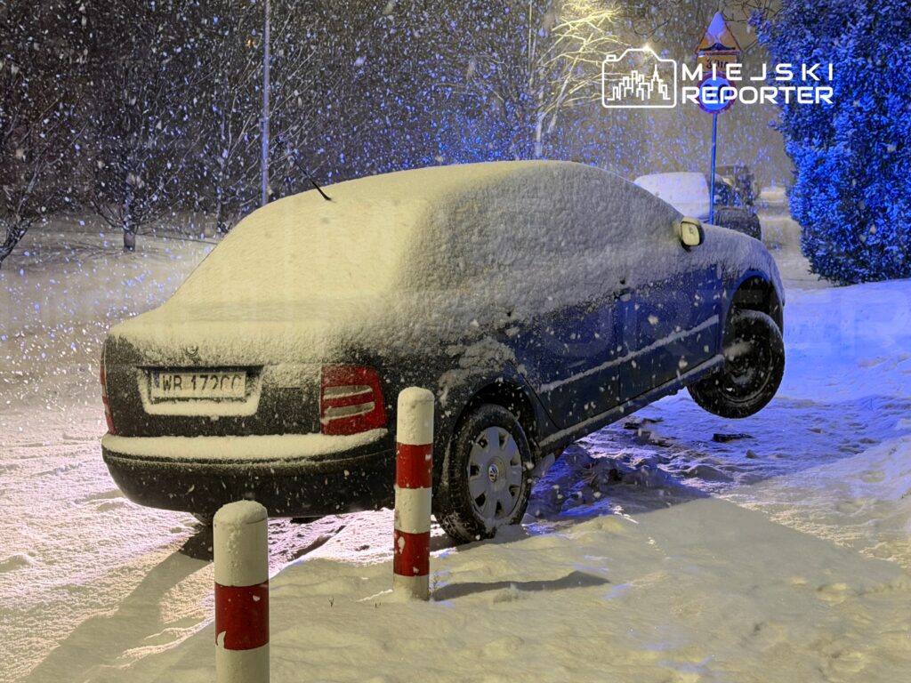 Samochód uniesiony na jednym kole, pokryty śniegiem, stoi na ośnieżonym chodniku w trakcie intensywnej śnieżycy.
