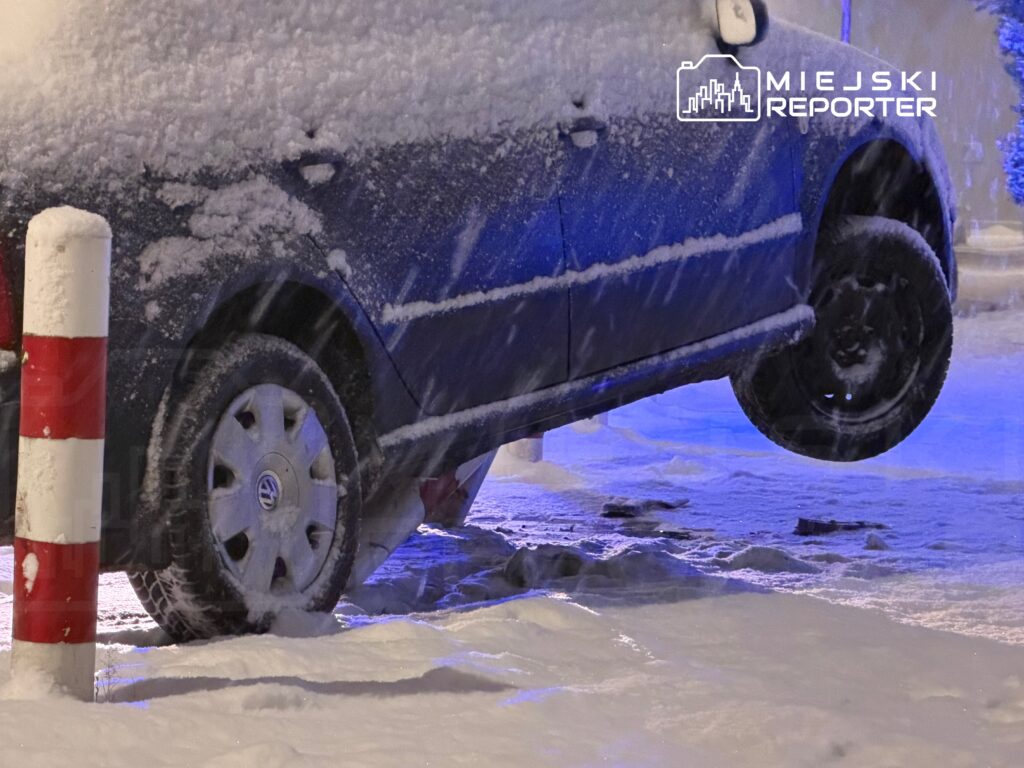 Samochód uniesiony na jednym kole w zasypanym śniegiem parkingu, z widocznymi opadami śniegu.
