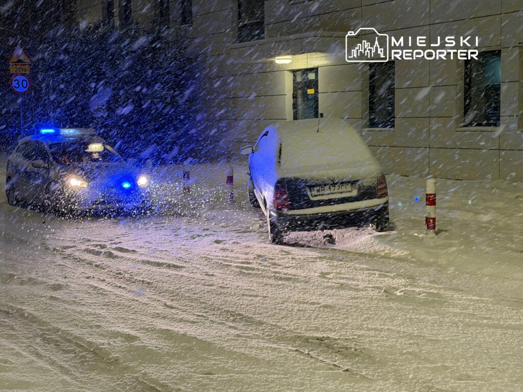 Policyjny radiowóz z włączonymi sygnałami świetlnymi stoi na zaśnieżonej ulicy obok zaparkowanego samochodu.