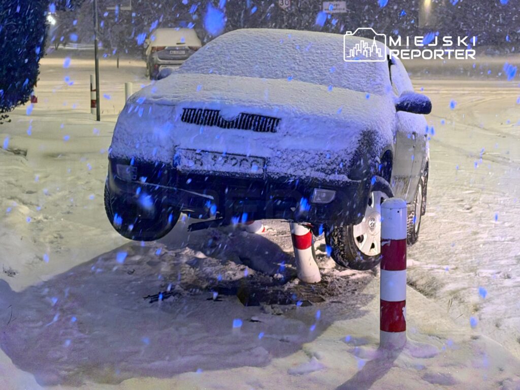 Samochód uniesiony nad ziemią, pokryty warstwą śniegu, stoi obok czerwono-białych słupków w zimowym otoczeniu.