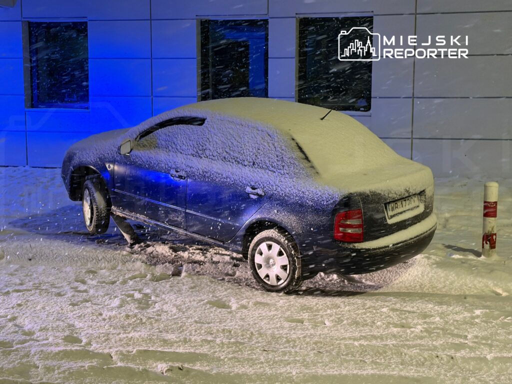 Niebieski samochód osobowy pokryty śniegiem stoi na zaśnieżonym parkingu w nocy, w tle widoczna ściana budynku.